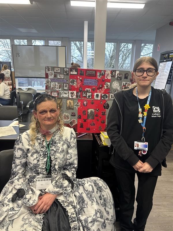 Two blonde ladies look at the camera. One is dressed up in a white and blue dress and the other is all in black wearing glasses. Behind them is a red mood board with ideas surrounding the competition.